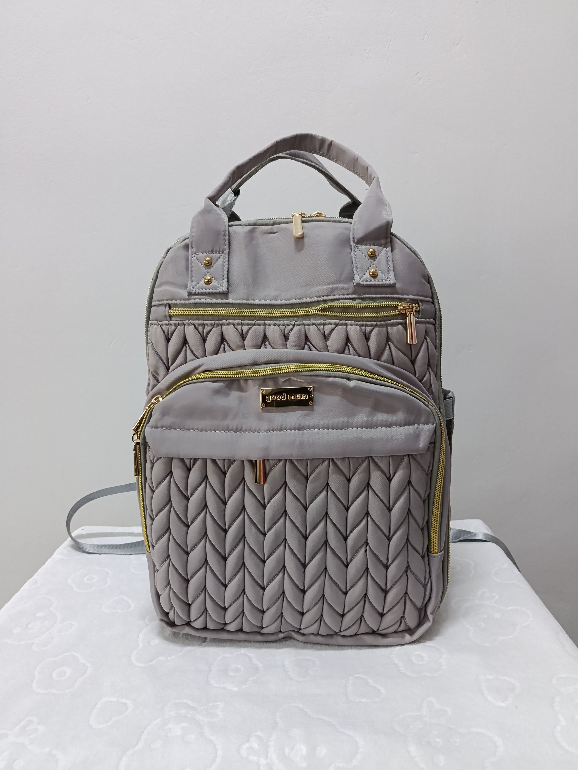 A gray quilted backpack with gold-tone zippers and hardware, featuring top handles and a front pocket, displayed on a white surface against a light gray background.