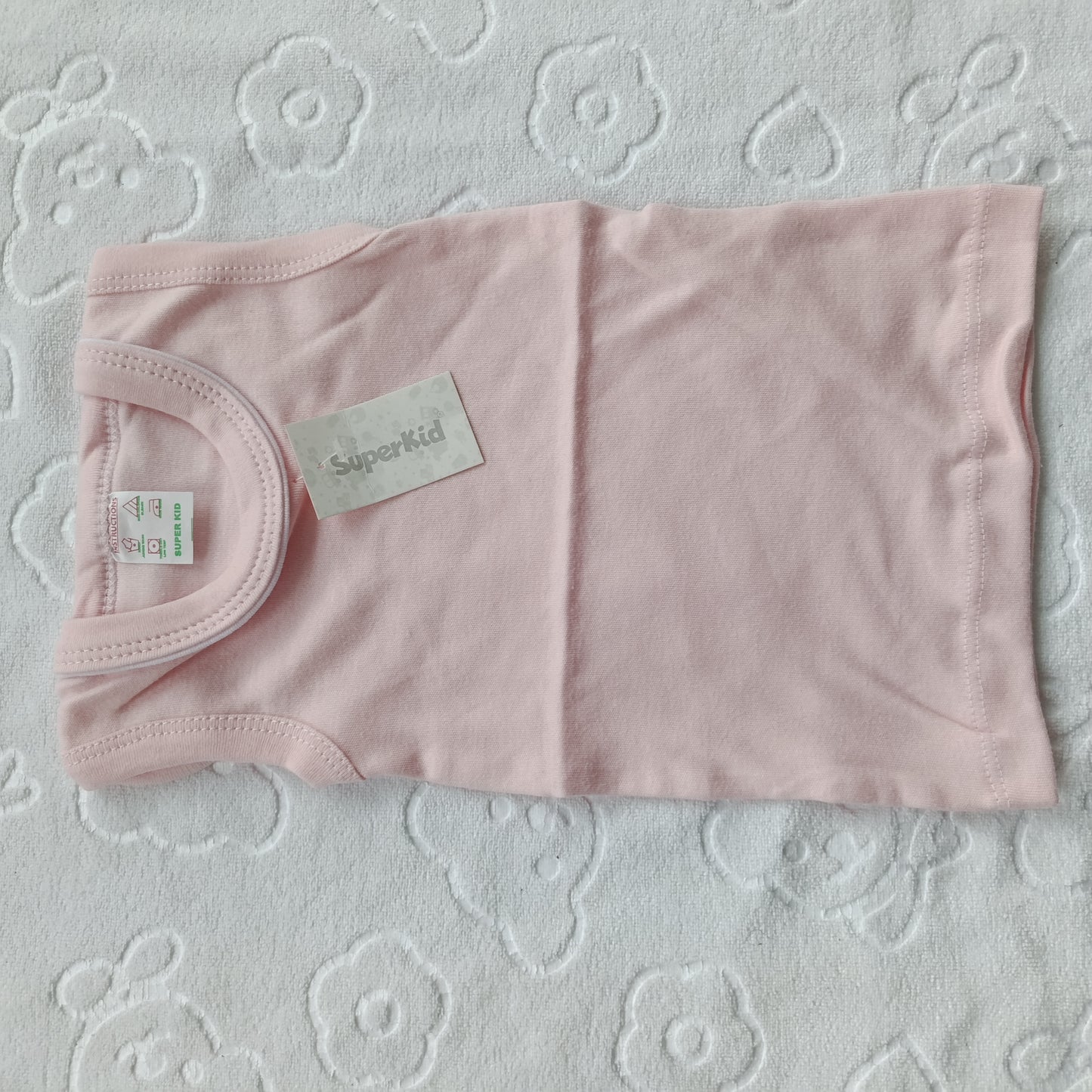 A light pink baby tank top laid flat on a white textured blanket with a floral pattern. The shirt has a visible 'Superkid' brand label and a size tag.