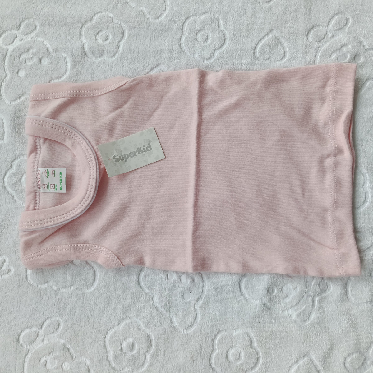 A light pink baby tank top laid flat on a white textured blanket with a floral pattern. The shirt has a visible 'Superkid' brand label and a size tag.
