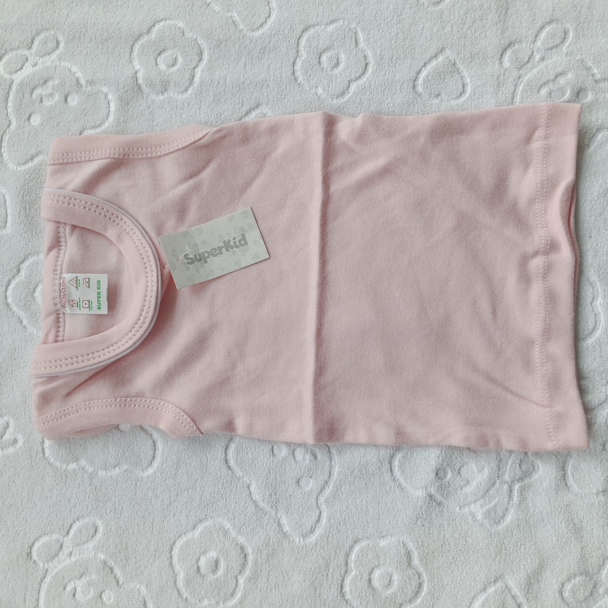 A light pink baby tank top laid flat on a white textured blanket with a floral pattern. The shirt has a visible 'Superkid' brand label and a size tag.